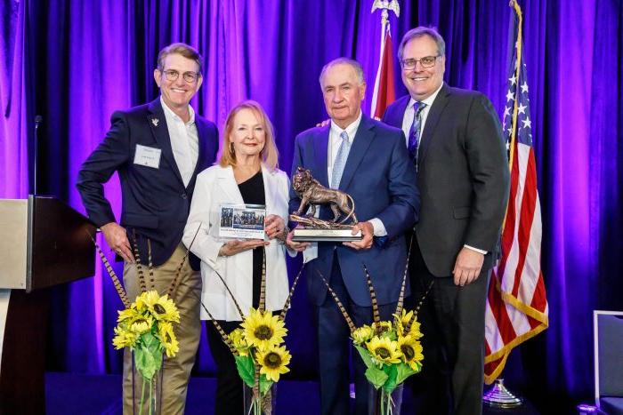 From left to right, Dr. Rudin, Senator Ted Lyon, Donna Lyon and Dr. 凯利·雷纳站在紫色幕布前的舞台上.S. and Texas flags. The Lyons are holding a lion statue. 在人们面前，地板上放着三束向日葵.